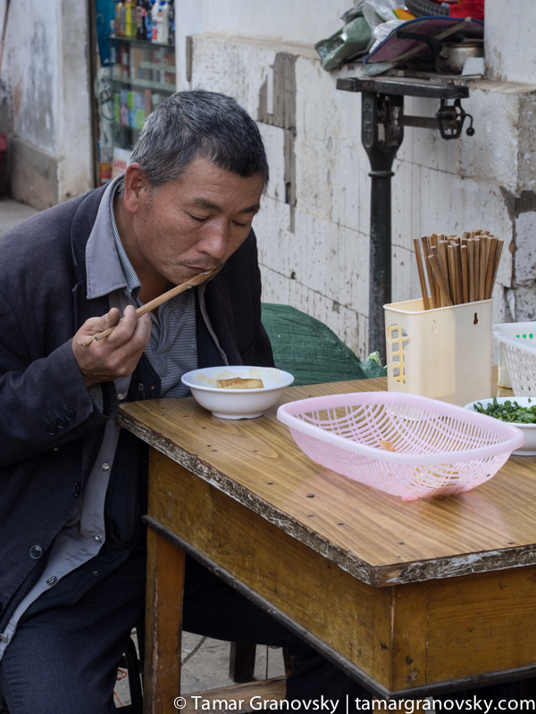A Man Sits and Eats Doufu and Rice Sticks for Breakfast at the Table Across from Me. I Do the Same.